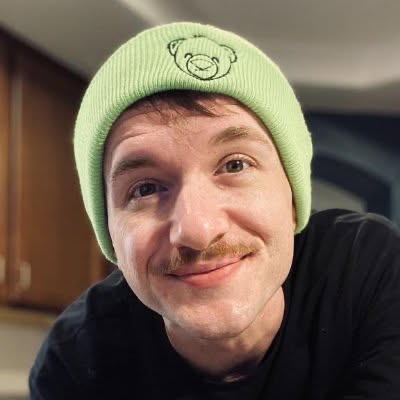 Close-up portrait of Cash in a green knit beanie with a small bear icon. Relaxed, direct, smiling.