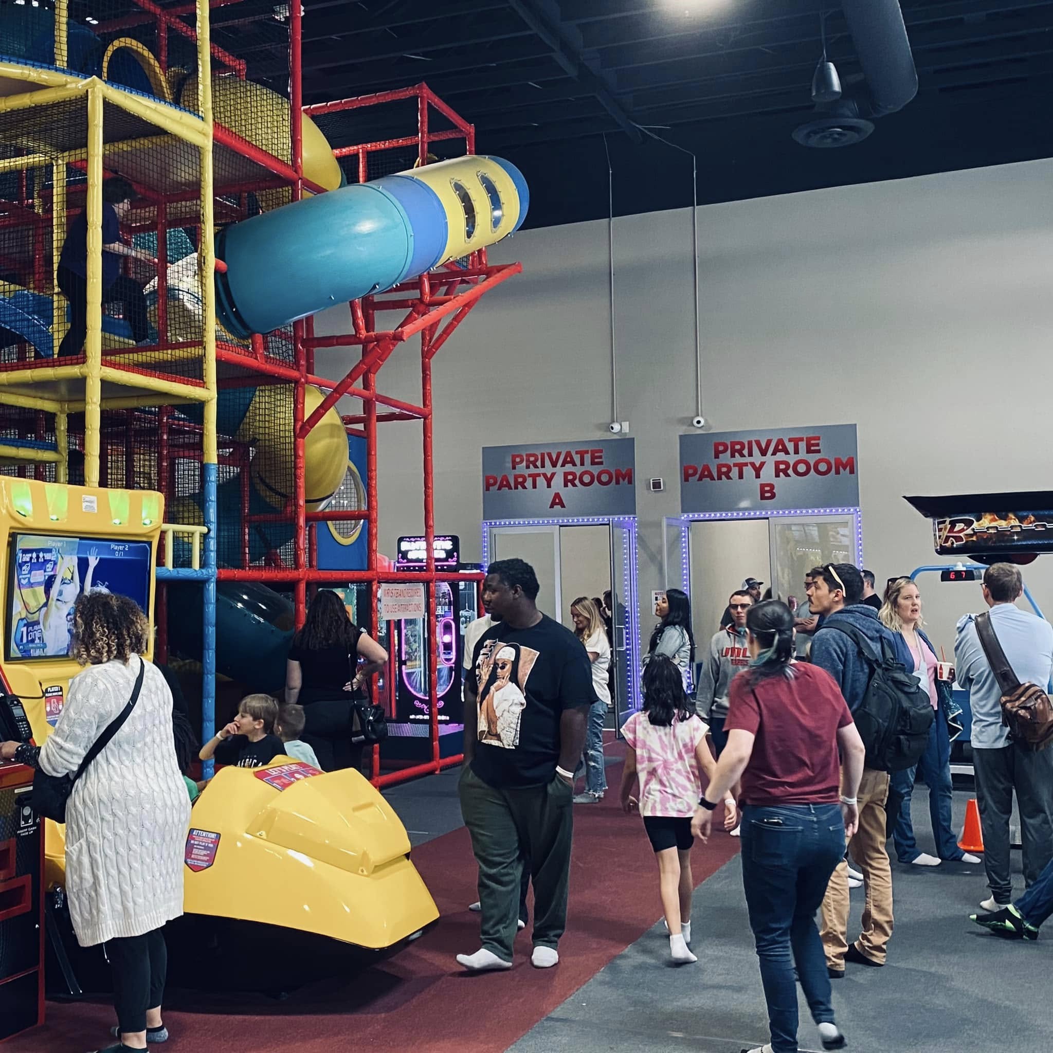 Inside a colorful indoor play center with a large multi-level play structure, slides, and party-room signage. Families and kids moving through the space.